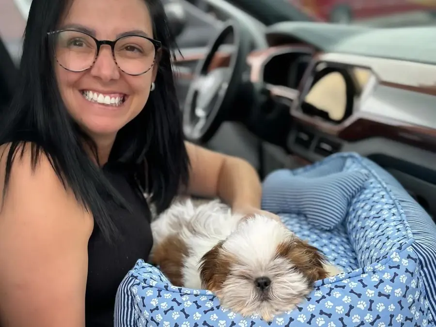 Linda from Tennessee smiling in her car with Sam, a gold and white Shih Tzu puppy, asleep in a blue bed on her lap